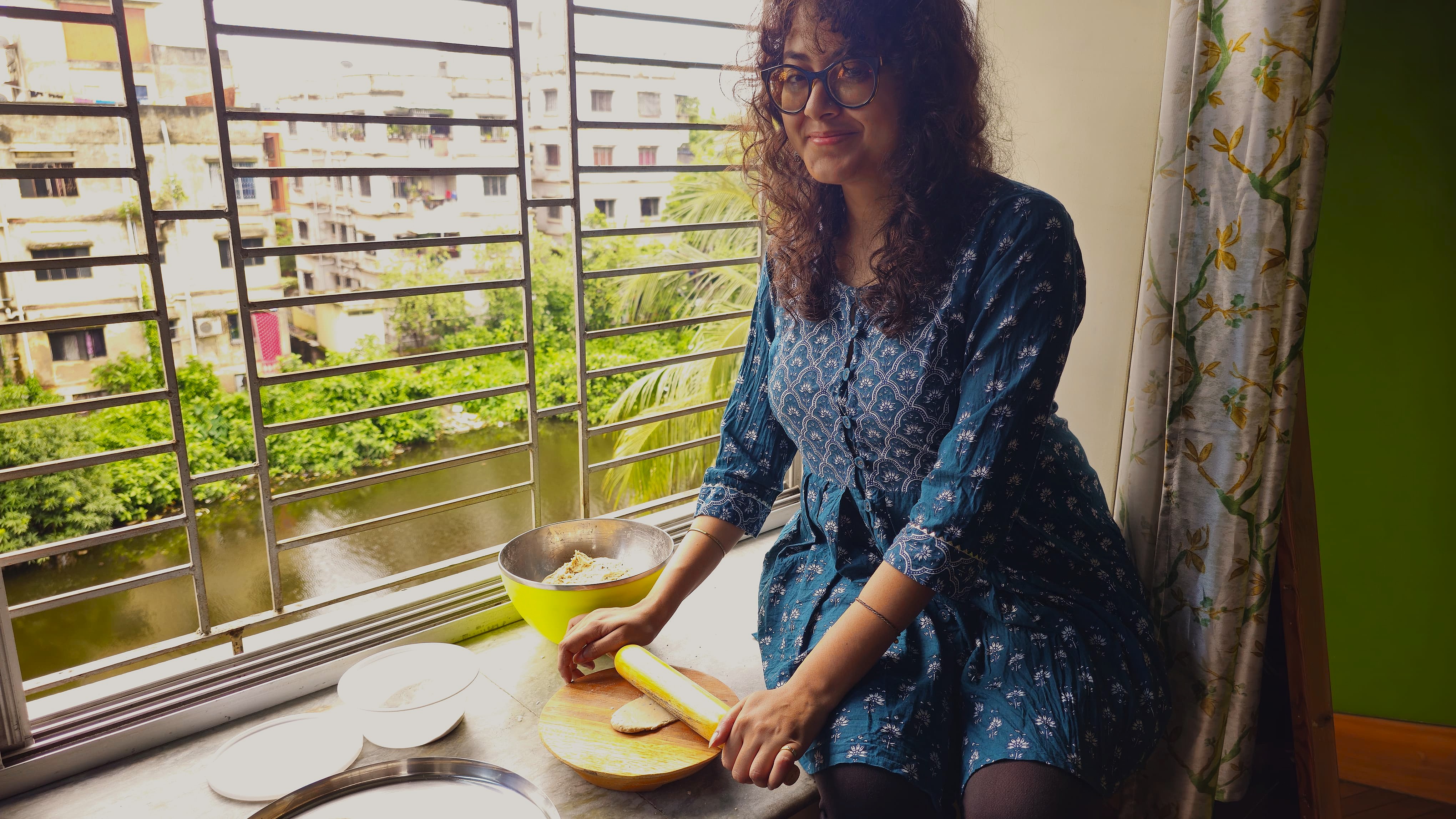 A person sitting near a window, rolling dough on a wooden board, with a bowl of ingredients and plates nearby, surrounded by a lush green view outside.