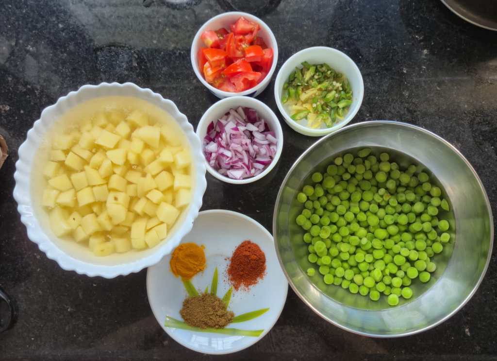 Five bowls and one small plate containing ingredients such as potatoes, tomatoes, garlic, ginger, chilies, green peas, onions, and spices.